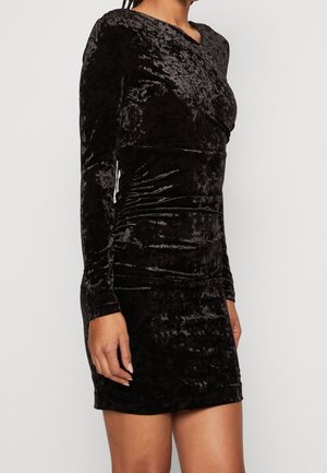 Kobieta ubrana w dopasowaną czarną mini sukienkę z długim rękawem z marszczonego aksamitu, z lekkim marszczeniem w talii i poduszkami na ramionach.