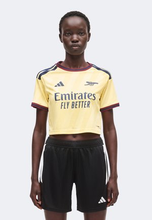 Νεαρό άτομο με κίτρινη κοντή φανέλα ποδοσφαίρου της Arsenal και μαύρο σορτς Adidas, στέκεται μπροστά σε απλό φωτεινό φόντο.