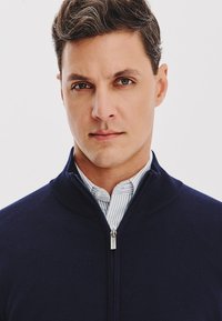 Granatowy sweter na zamek wykonany z miękkiego materiału dzianinowego, z wysokim kołnierzem i szczotkowanym wykończeniem. Nosi się go na jasnoniebieskiej, w paski koszuli.