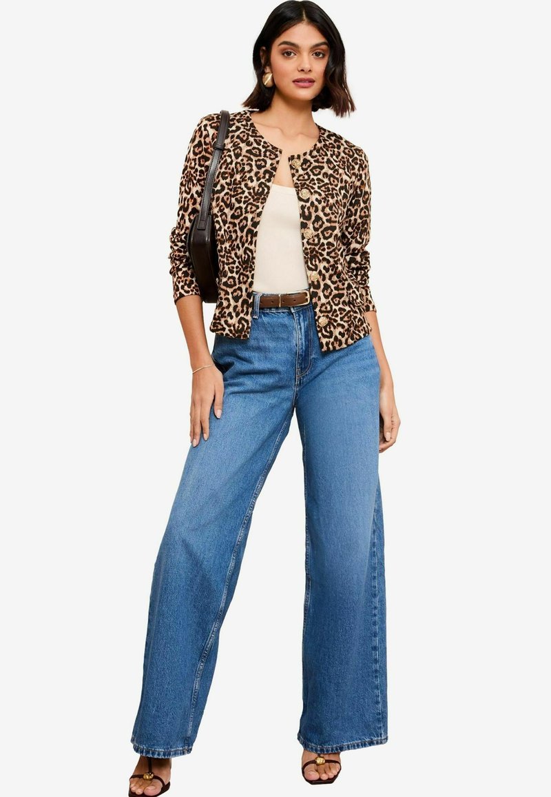 Cardigan leopardato, aderente, con scollo rotondo, abbinato a jeans in denim a vita alta e gamba larga. Il modello indossa una maglietta beige e accessori.