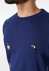 Sudadera de color azul marino hecha de tela suave que presenta diseños bordados de un tucán, una palmera y un coco con pajitas.