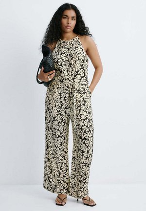 Mujer con cabello largo y rizado, vestida con un mono sin mangas floral beige y negro, sosteniendo un bolso tejido negro, de pie con una mano en el bolsillo.
