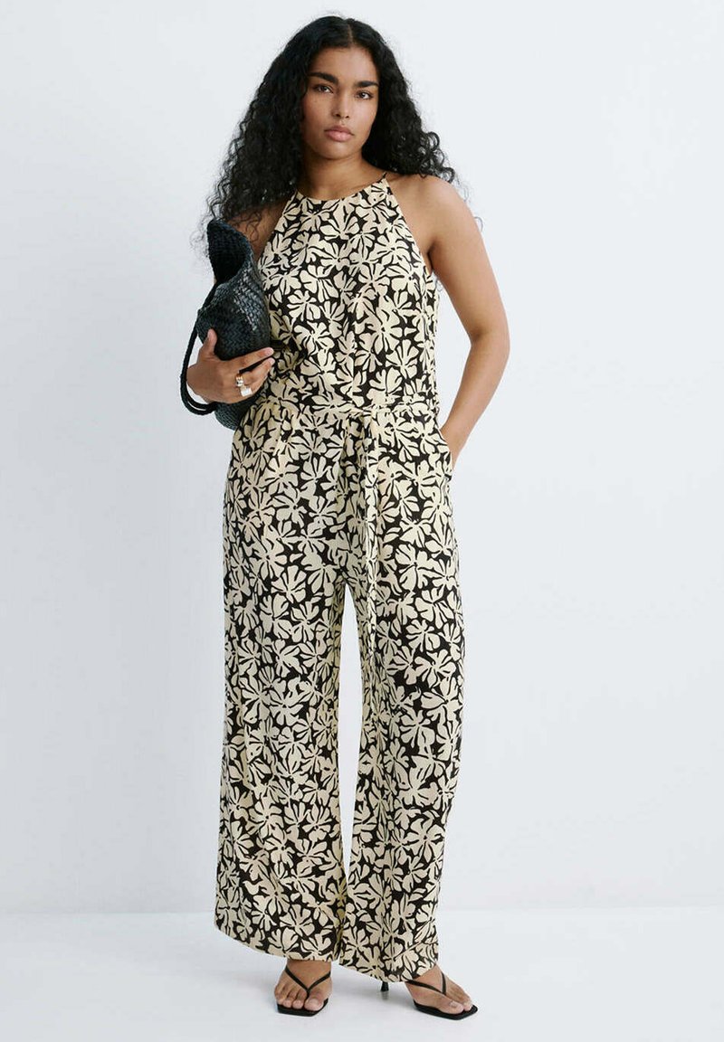 Femme aux longs cheveux bouclés portant une combinaison beige et noire à fleurs sans manches, tenant un sac noir tissé, debout avec une main dans la poche.