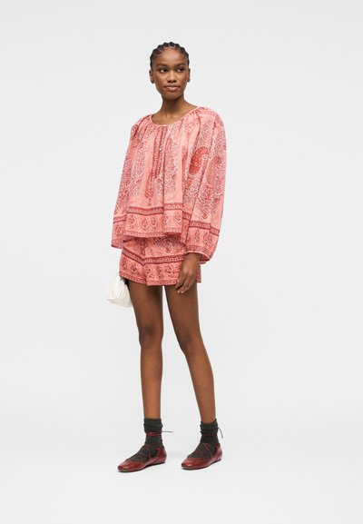 Femme portant un chemisier ample à motifs roses et un short assorti, tenant un petit sac blanc, portant des chaussettes foncées et des ballerines rouges, debout sur un fond blanc.
