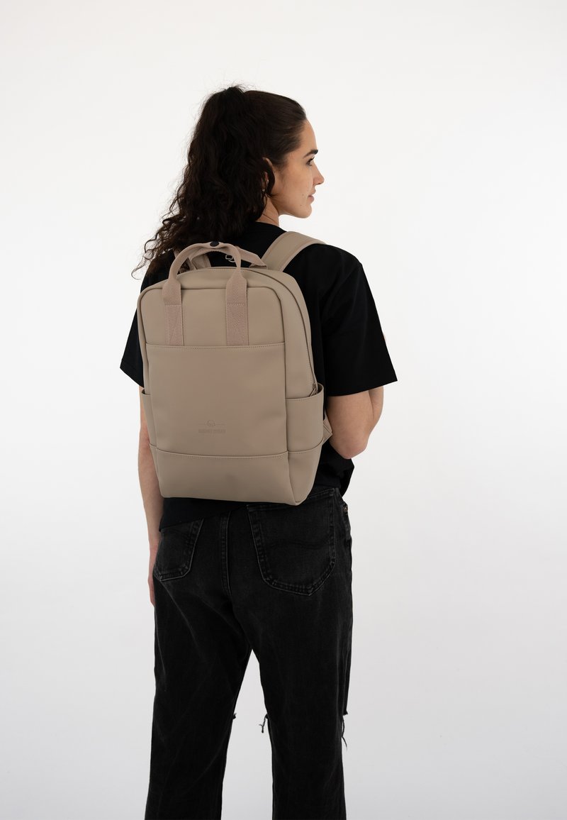 Beige backpack made of soft material with dual top handles and padded straps, featuring a front pocket and subtle logo detail.
