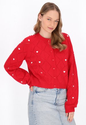 Punainen neulecardigan, jossa on timanttikuviot ja pieniä valkoisia sydämiä, ribbireunalla kauluksessa ja hihoissa, kiinnitetty vastaavilla napeilla.