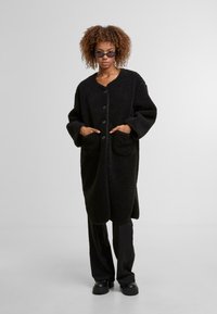 Cappotto lungo in pile nero con scollo a V, chiusura a bottoni, grandi tasche frontali e maniche larghe. Indossato sopra pantaloni neri larghi e scarpe chunky.