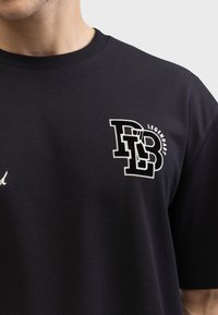 Černé bavlněné tričko s bílým grafickým logem a textem "PB" a "LEGENDARY". Materiál má hladkou texturu a kulatý výstřih.