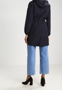 Osoba oblečená v černém kapucovém kabátu, světle modrých zkrácených džínách a černých botách na podpatku, stojící zády k divákovi.