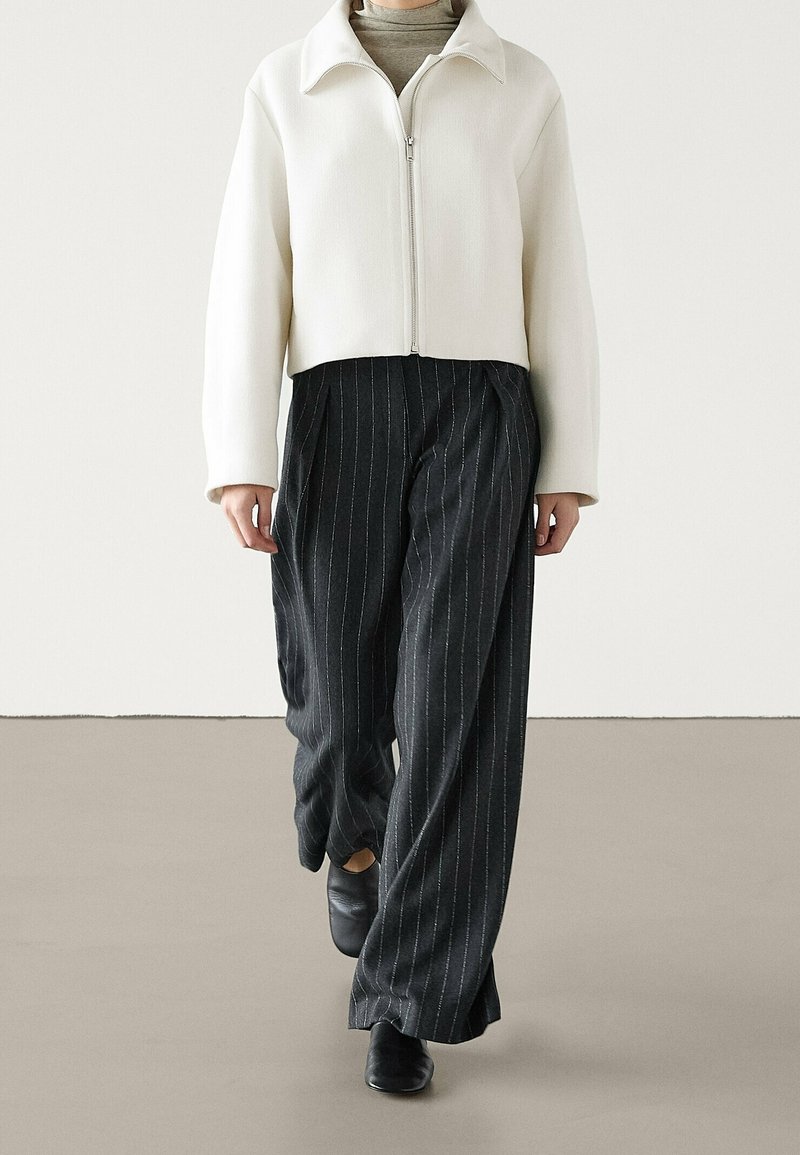 White cropped jacket with a high collar, zip closure, paired with black pinstriped wide-leg pants and black shoes.