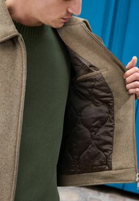 Braune, strukturierte Außenjacke mit einer gesteppten, dunkelbraunen Fütterung und einer Seitentasche. Darunter ein grüner Pullover, der Reißverschluss ist sichtbar.