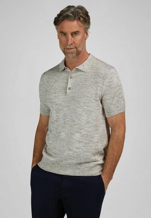 Hombre con cabello y barba gris, vestido con una camisa polo de manga corta gris claro y pantalones oscuros, de pie con las manos en los bolsillos contra un fondo liso.