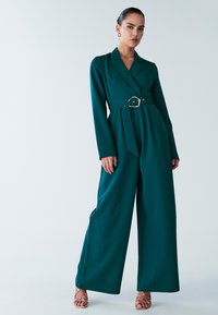 Green, long-sleeve jumpsuit with a wrap design, belted waist featuring a gold buckle, and wide trousers. Smooth fabric with no patterns.