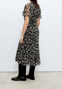 Vestido midi negro con estampado floral y mangas cortas transparentes, que presenta una cintura ajustada y una falda fluida. Combinado con botas negras hasta la rodilla.