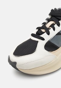 Gros plan d'une chaussure de sport blanche et noire avec des lacets noirs, un tissu noir texturé au niveau des orteils et une semelle intermédiaire beige sur un fond clair.