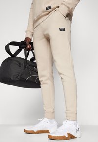 Beige bomull joggebukser med sidelommer og svarte etiketter, kombinert med hvite joggesko med teksturdesign og såler av gummi. Svart treningsbag.