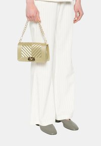 Borsa a tracolla dorata trapuntata con catena, caratterizzata da una forma rettangolare, un motivo orizzontale e un gancio metallico. Indossata con pantaloni larghi a righe bianche.