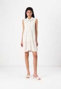 Robe sans manches de couleur crème avec un col, un devant boutonné et une jupe à volants. Portée avec des mules à talons de couleur claire.