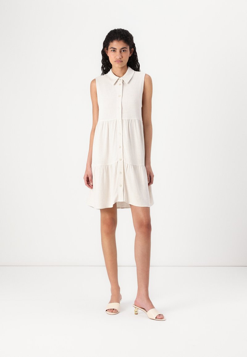 Robe sans manches de couleur crème avec un col, un devant boutonné et une jupe à volants. Portée avec des mules à talons de couleur claire.
