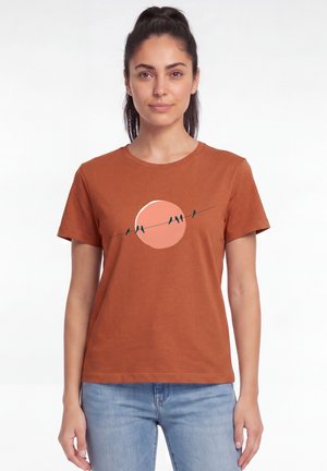 Vrouw met donker haar die een roestkleurig T-shirt draagt met een vogel-silhouet ontwerp op een ronde perzikkleurige achtergrond, gecombineerd met lichtblauwe spijkerbroek.