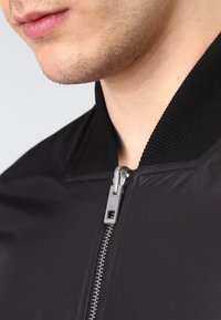 Svart bomberjacka med en ribbad krage och en silvrig dragkedja. Tyget ser slätt ut med en lätt textur.