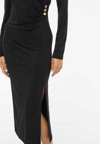 Juoda ilgomis rankovėmis suknelė su krintančiu dizainu, šoniniu plyšiu ir trim auksiniais mygtukais kairėje pusėje. Pagaminta iš lygaus audinio.