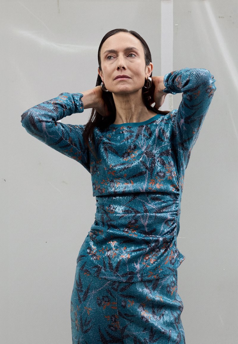 Robe turquoise ornée de paillettes avec manches longues et motif floral. Le tissu présente un effet scintillant et un léger fronces à la taille, encolure ronde.