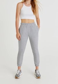 Graue Jogginghose mit elastischem Bund und Seitentaschen, kombiniert mit einem weißen, kurzen Tanktop und grauen Sportschuhen.