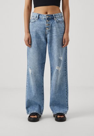 Femme portant un jean bleu taille haute à jambes larges avec des boutons, des patchs usés, des sandales noires et un top court noir sur un fond uni.