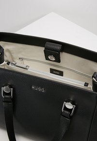 Czarna skórzana torebka Hugo z srebrnymi okuciami, otwarta u góry, ukazująca beżowe wnętrze z kieszeniami na zamek.