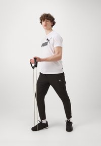 Puma træningstøj: hvid t-shirt med sort logo, sorte cargo-bukser, holder i modstandsbånd, sorte sportssko, hvide sokker, krøllet hår.