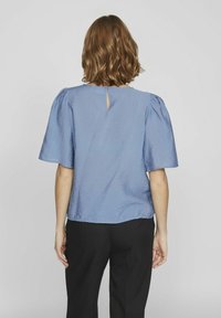 Haut bleu clair avec des manches courtes bouffantes, en textile texturé, et un détail en forme de clé au dos, associé à un pantalon noir.