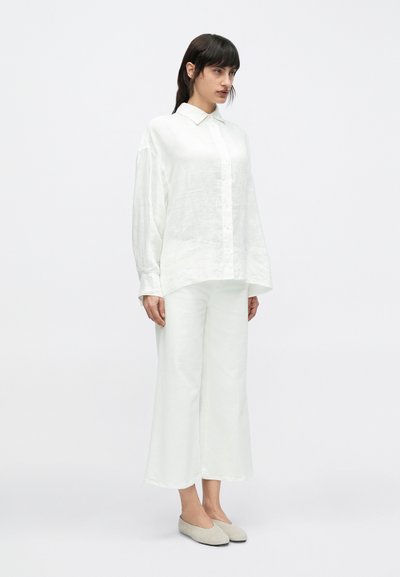 Λευκό πουκάμισο από λινό με κουμπιά και γιακά, συνδυασμένο με λευκό παντελόνι φαρδιάς γραμμής. Το μοντέλο φοράει ανοιχτό γκρι επίπεδα παπούτσια. Μινιμαλιστικός σχεδιασμός.
