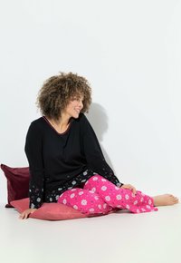 Černý top s dlouhými rukávy s akcenty hvězdného vzoru a růžové pyžamové kalhoty se bílými květinovými vzory. Sedí na růžovém polštáři.