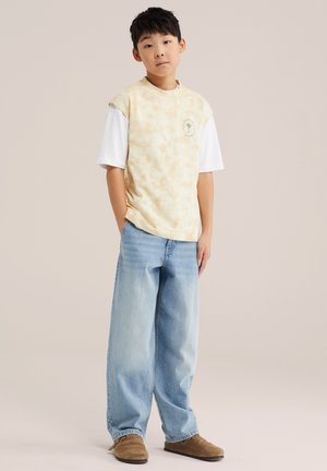 Noor inimene lühikeste mustade juustega, kes kannab beeži tie-dye mustriga varrukateta särke valge T-särgi peal, laia hele sinist teksapükse ja pruune kingi, seistes.