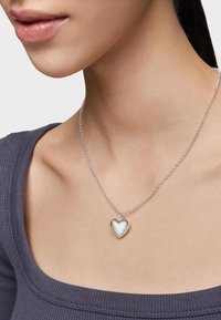 Collar de péndulo en forma de corazón de plata sobre una delicada cadena, llevado en un cuello desnudo. El corazón tiene una superficie lisa y reflectante y está unido a una cadena simple.