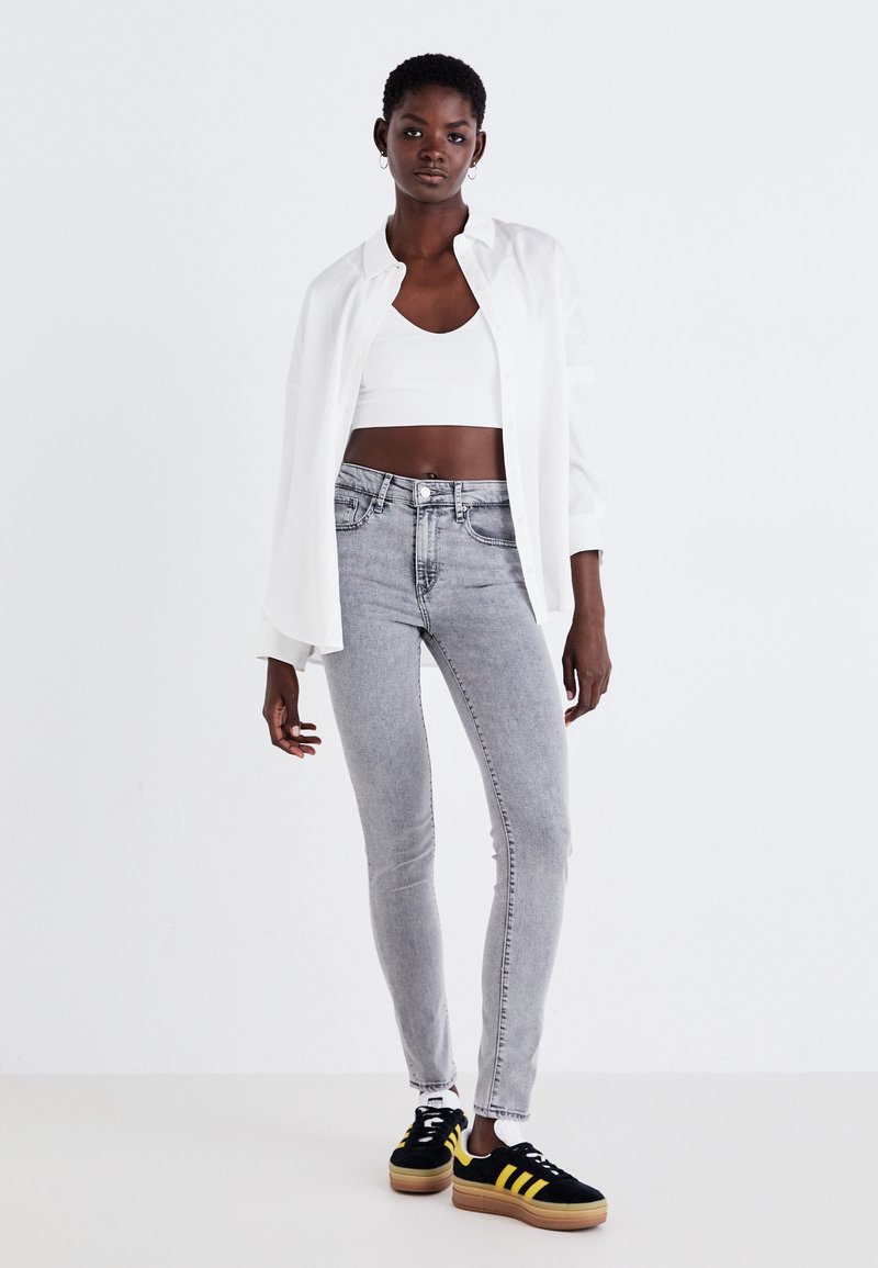 Weißes Hemd über einem weißen Cropped-Top, kombiniert mit hellgrauen Skinny-Jeans und schwarzen Sneakers mit gelben Streifen. Neutraler Hintergrund.