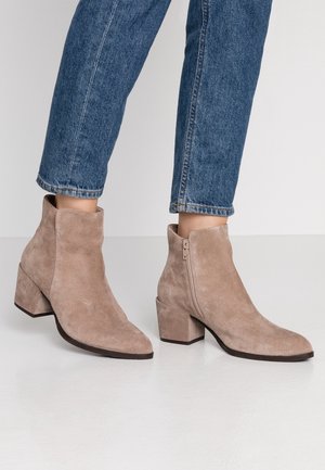 Personne portant des bottines en daim beige à talons blocs et fermetures éclair latérales, associées à un jean bleu denim sur un fond uni.