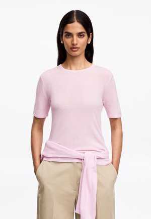 Femme aux longs cheveux foncés porte un haut rose clair à manches courtes et un pantalon beige avec un pull rose assorti noué autour de la taille sur fond blanc.