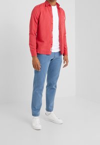 Rode button-up overhemd over een wit T-shirt, gecombineerd met lichtblauwe jeans en witte sneakers. Soepele stof, klassieke kraag en een ontspannen pasvorm.