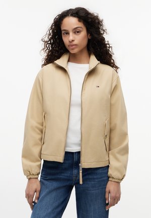 Kvinde med krøllet hår iført en beige lynlåstrøje over en hvid skjorte og blå jeans, stående mod en ensfarvet hvid baggrund.
