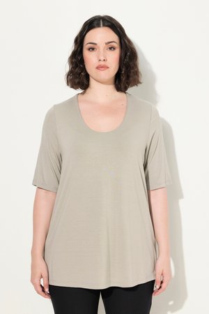 Frau mit schulterlangem dunklem Haar, die ein beiges kurzärmliges Top mit Rundhalsausschnitt und schwarze Hose trägt, vor einem weißen Hintergrund stehend.
