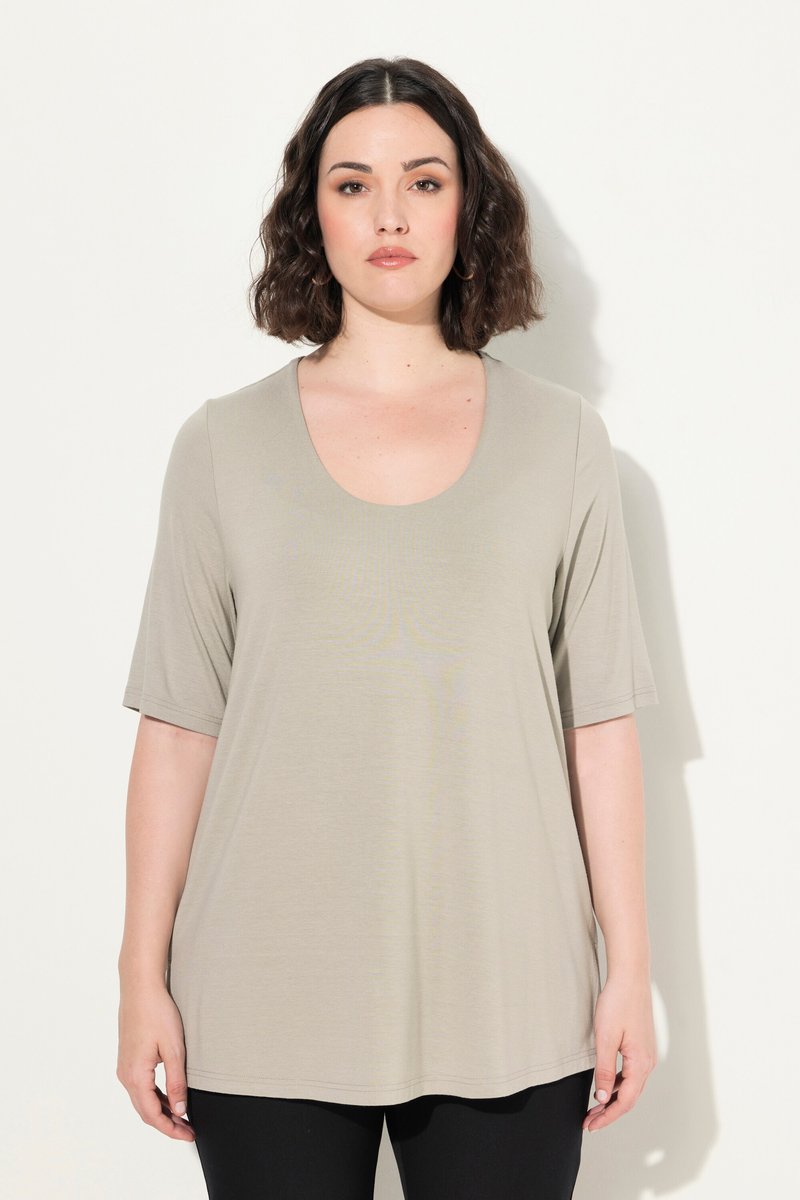 Vrouw met donker haar tot op de schouders, gekleed in een beige top met korte mouwen en ronde hals en zwarte broek, staand tegen een witte achtergrond.