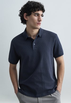 Μπλε ναυτικό πόλο μπλουζάκι με υφασμένο σχέδιο σε σχήμα chevron, κοντά μανίκια και γιακά. Διαθέτει κούμπωμα με κουμπιά και άνετη γραμμή.