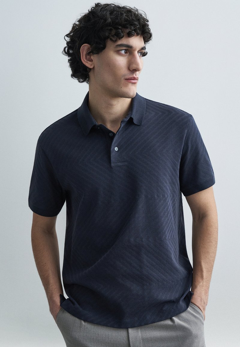 Tumši zils polo krekls ar fakturētu zigzaga rakstu, īsām piedurknēm un apkakli. Ir pogu aizdare un brīvs piegriezums.