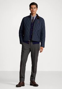 Marinblå vadderad jacka med bruna detaljer, bärs över en marinblå cardigan, grå byxor och bruna läderskor; har dragkedja och fickor.