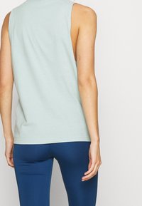 Top verde chiaro senza maniche con una texture liscia, caratterizzato da una scollatura rotonda e ascellari aperti, abbinato a leggings aderenti blu scuro.