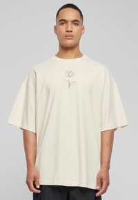 Kremfarget oversized t-skjorte laget av myk bomull, med et svart linjete rose-design i midten. Korte ermer og rund hals.