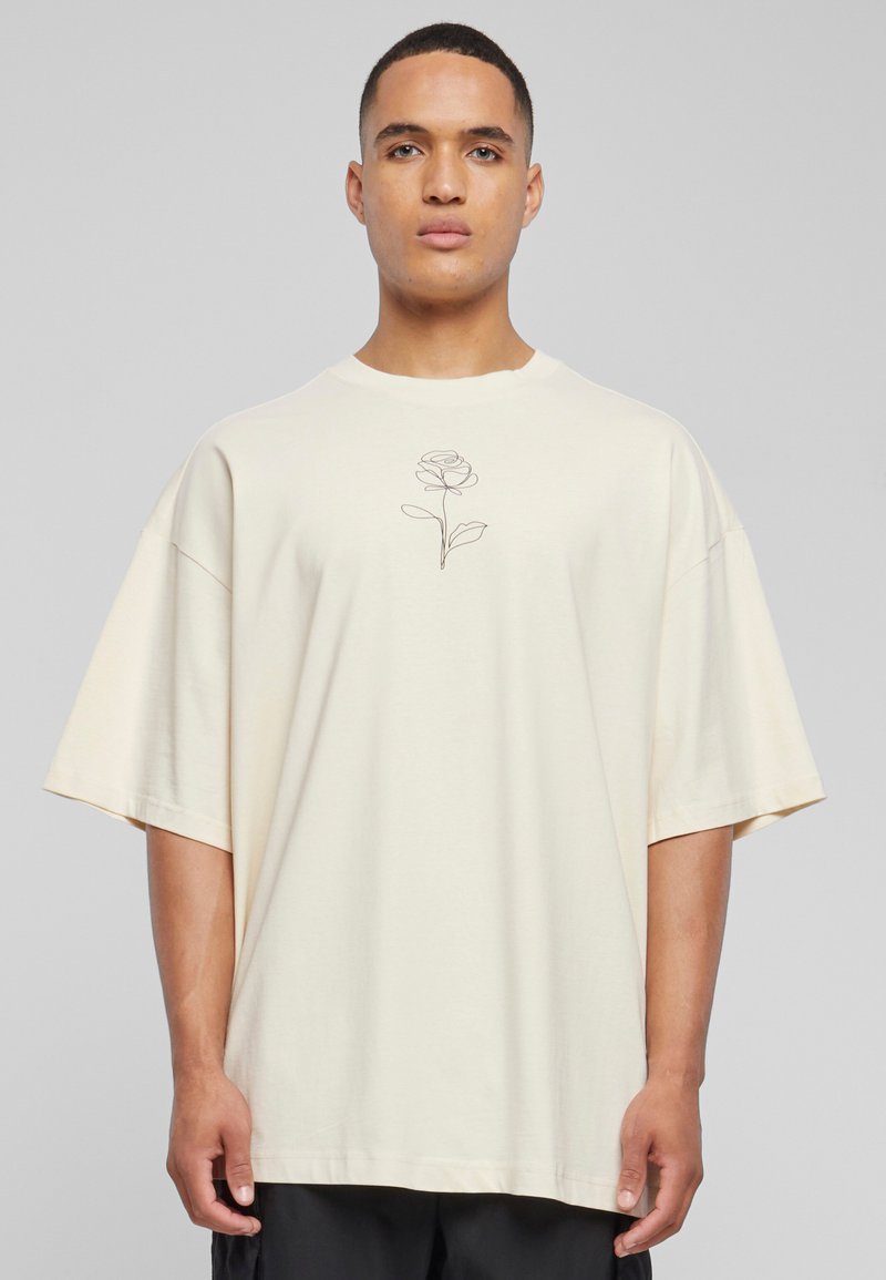 Kremfarget oversized t-skjorte laget av myk bomull, med et svart linjete rose-design i midten. Korte ermer og rund hals.
