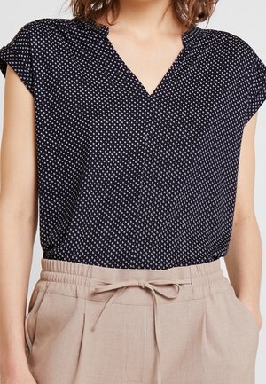 Frau trägt eine schwarze Bluse mit kurzen Ärmeln und kleinem weißem Muster sowie eine beige Hose mit hoher Taille und gebundenem Gürtel.